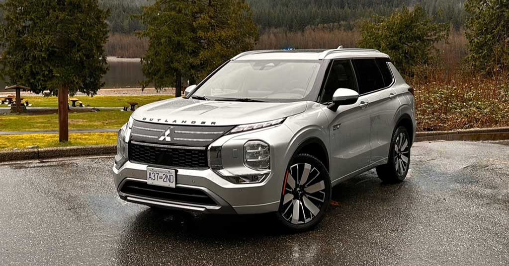 2026-Outlander-PHEV • Edmonton's Business Front-three-quarter view of a silver Mitsubishi Outlander SUV parked on a wet pavement with trees and a lake in the background, daytime outdoor scene.